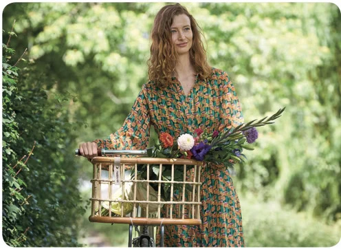 Woman in a patterned dress standing beside her bicycle with a basket full of colorful flowers, set in a lush green outdoor environment.