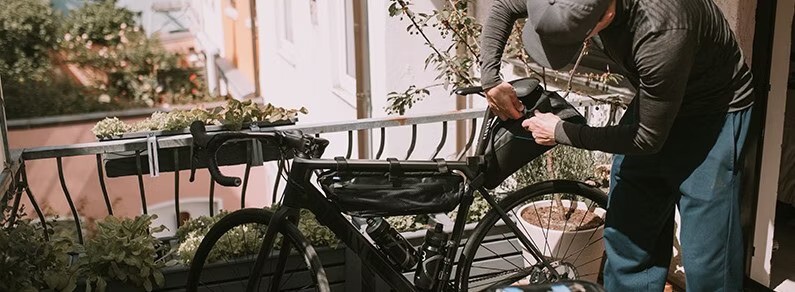 Person adjusting bikepacking bags on a bicycle on a balcony, surrounded by plants and an urban backdrop.