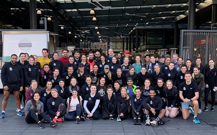 Large group of people in athletic wear posing outdoors under a covered walkway, with a mix of smiles and relaxed expressions.