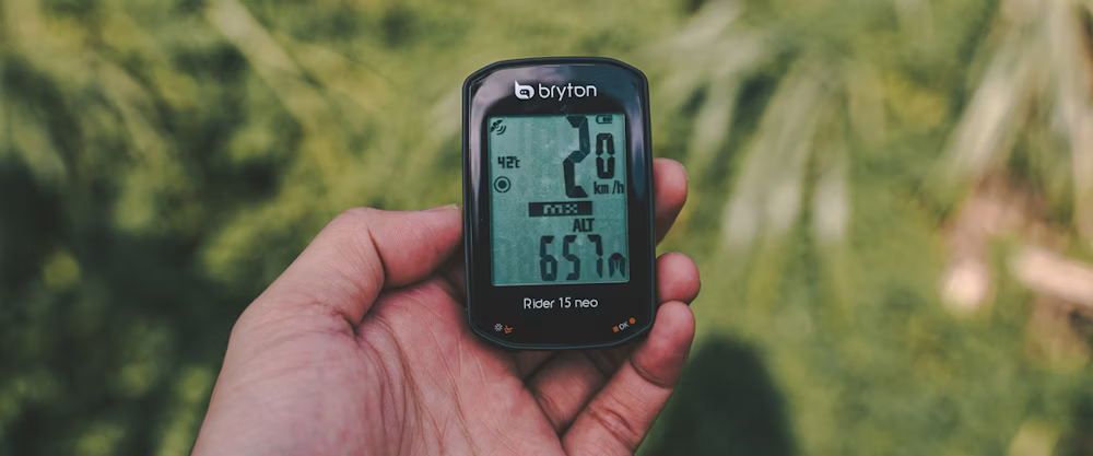 Hand holding a Bryton Rider 15 Neo cycling computer, displaying speed, altitude, and ride metrics.