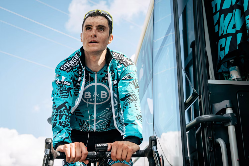Cyclist wearing a B&B Hotels team jersey on a stationary bike next to a team bus, with a bright sky in the background.