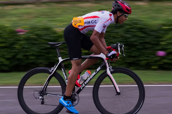 Action shot of a cyclist in a red helmet and white and red jersey riding a road bike at high speed on a smooth road, with a blurred green background.