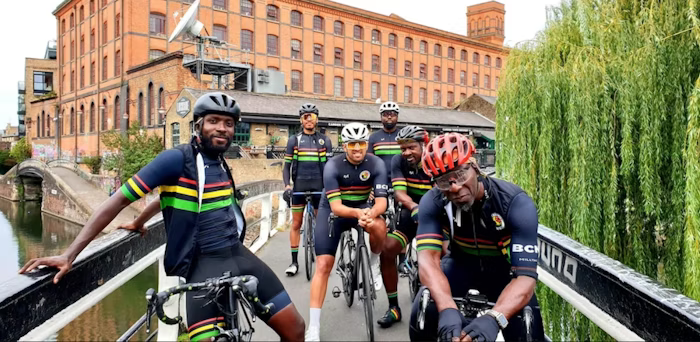 Group of cyclists in matching black kits with colorful stripes, posing on a bridge near an industrial building and canal, with greenery in the background.