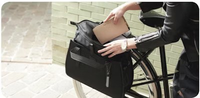 Person placing a notebook into a black bicycle bag attached to the rear rack of a bike, with a light green brick wall in the background.