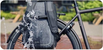 Close-up of a bicycle with a black waterproof backpack attached to the rear rack, water splashing onto the bag, with a blurred outdoor background.