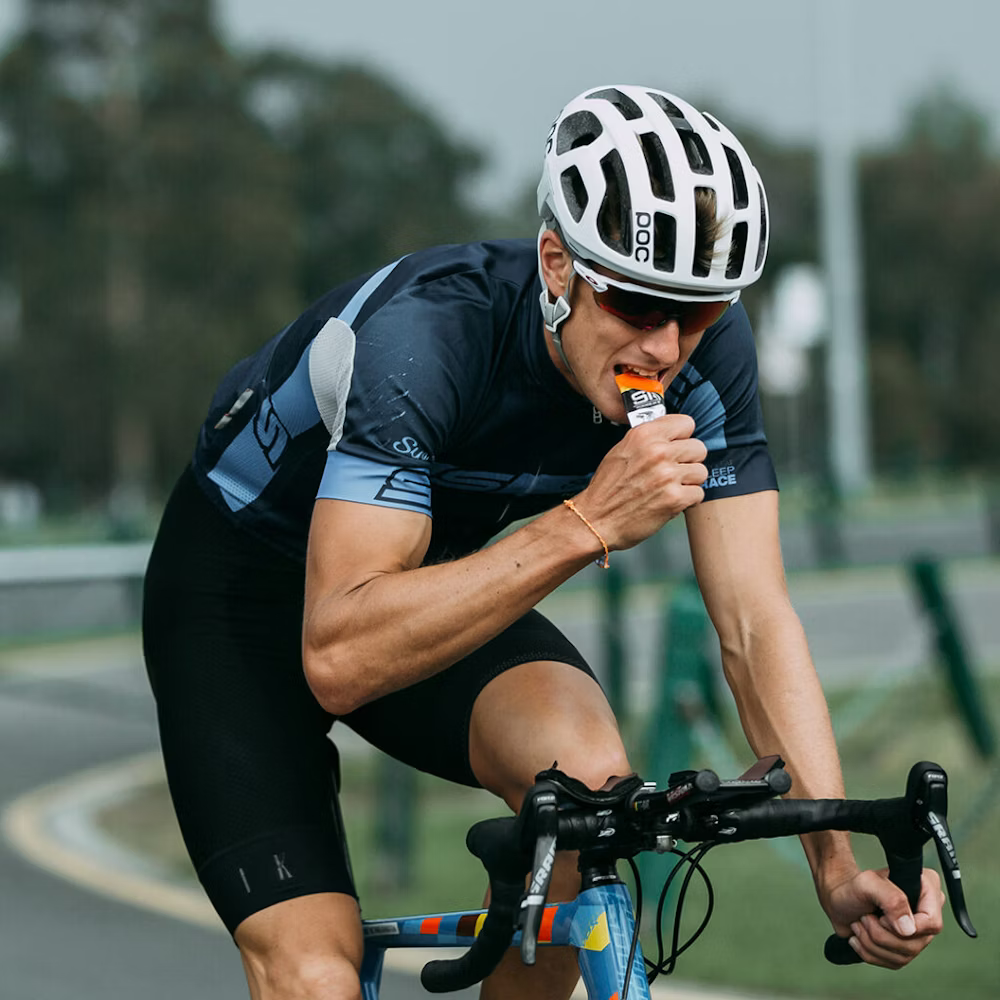 A cyclist in a white helmet and sunglasses eating an energy gel while riding a bike on a road.