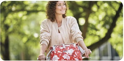Woman riding a bicycle with a red floral bag on the front rack, surrounded by greenery.