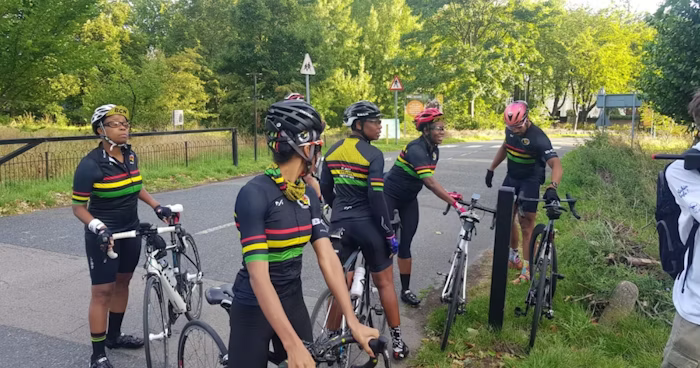 Group of cyclists wearing matching black kits with colorful stripes, gathered by the roadside with their bikes, surrounded by green trees and a rural setting.