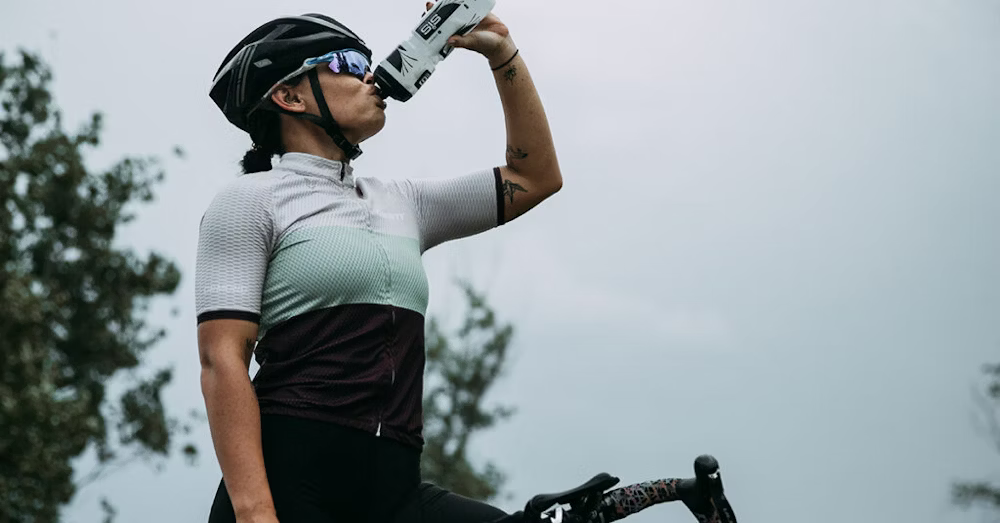 A cyclist in a helmet and sunglasses drinking from a water bottle while standing beside their bike on a cloudy day.