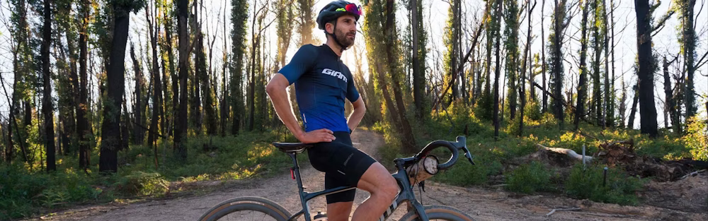 Cyclist wearing a blue and black kit with a helmet and sunglasses, standing next to a bike in a forest at sunset.