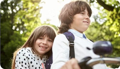 Two smiling children enjoying a ride on a bicycle in a sunny park setting.