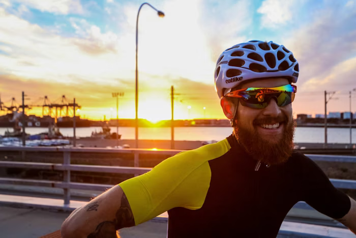 Cyclist wearing a white helmet and reflective sunglasses, smiling during a sunset at a harbor.