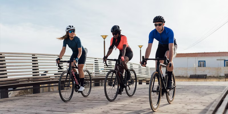 Three cyclists riding together on a wooden boardwalk, wearing cycling gear and helmets.