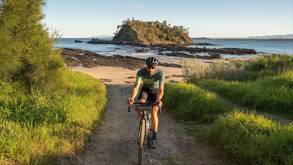 Cyclist riding on a grassy path near a scenic beach with an island in the background.
