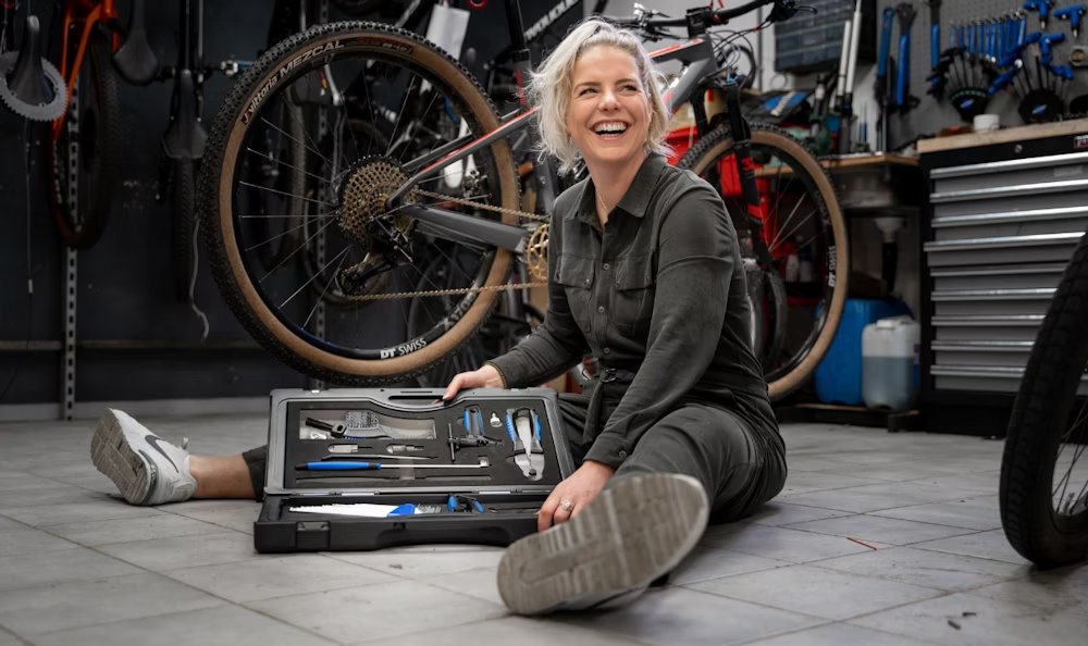 Smiling woman sitting on the floor of a bike workshop, holding a toolkit with bicycles in the background.