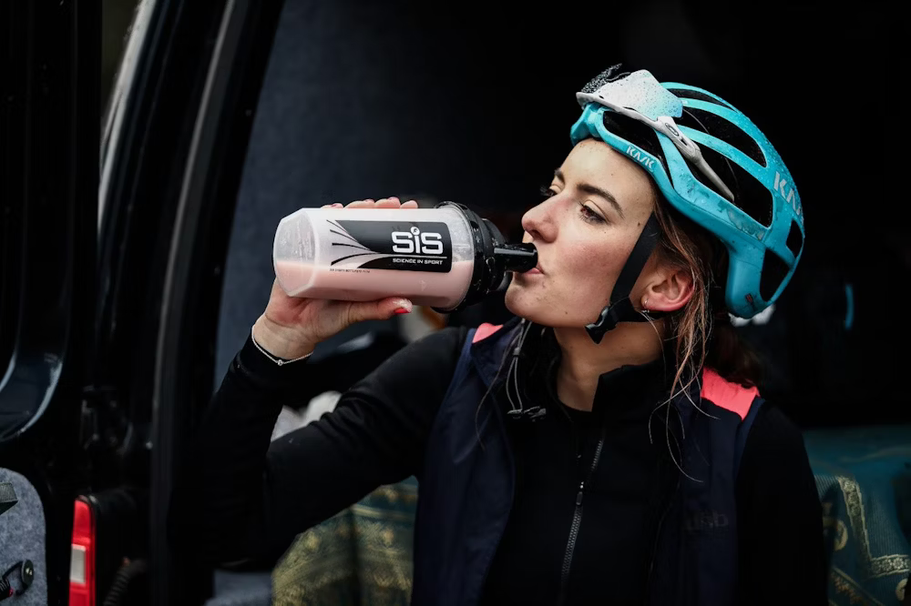 Cyclist wearing a blue helmet drinking from a SiS water bottle.