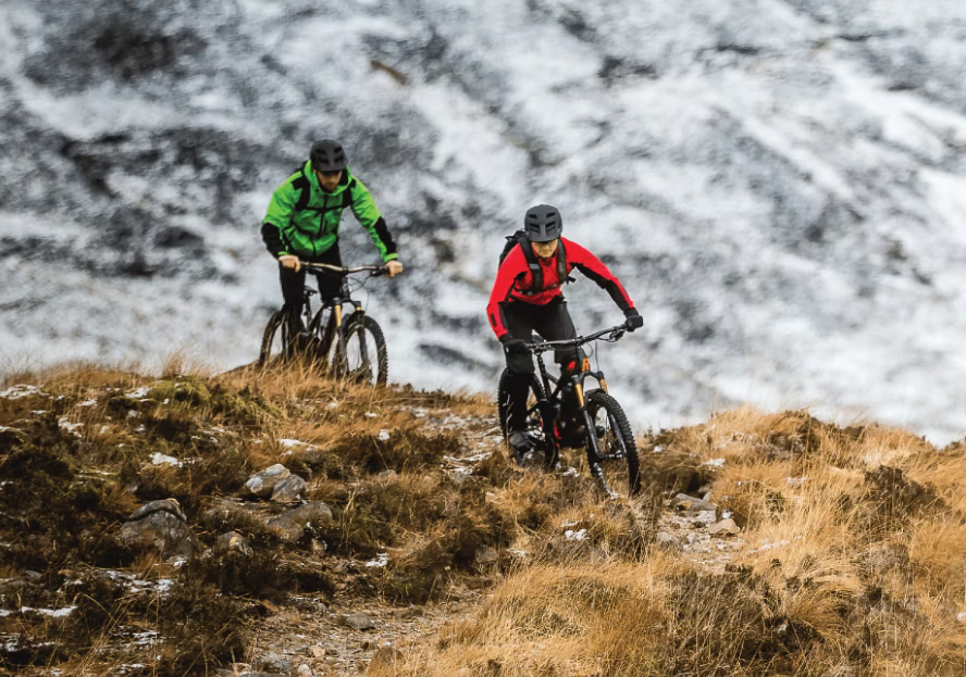 Two mountain bikers riding on a rugged trail in a mountainous area with snow in the background.