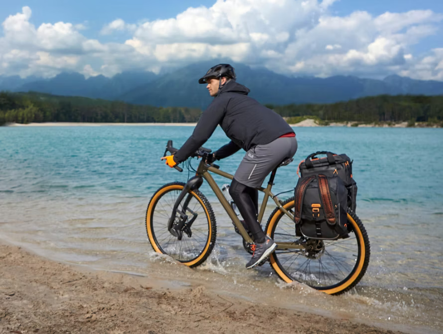 Cyclist riding a bike along the shore of a scenic lake with mountains in the background.