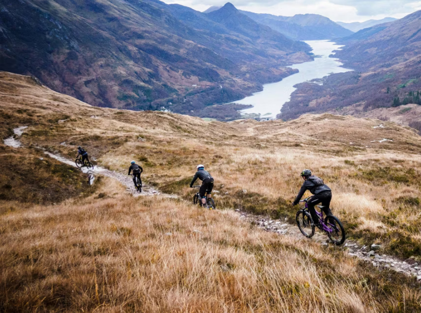 Group of cyclists riding down a mountain trail with a scenic valley and river in the background.