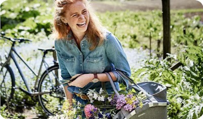 Woman smiling outdoors, carrying a basket filled with flowers, with a bicycle in the background.