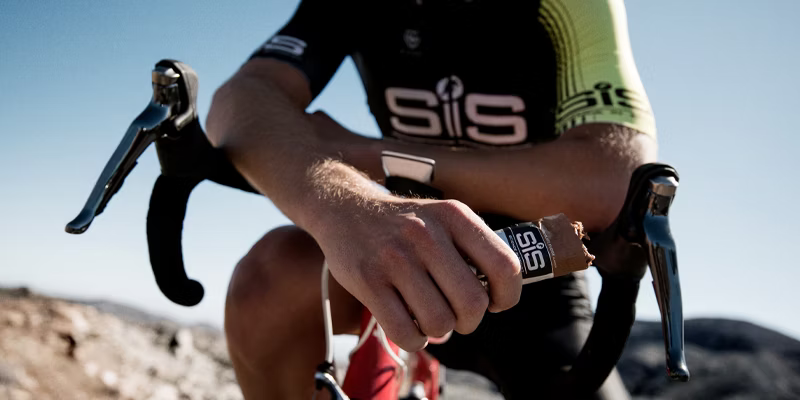 Cyclist sitting on a bike, holding an SiS energy bar, wearing a black and green cycling jersey.