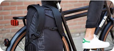 Close-up of a black backpack attached to a bicycle rack, with a person wearing white sneakers standing beside it.