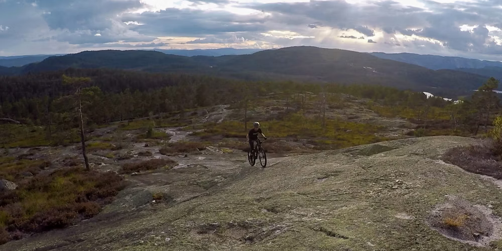 Cyclist riding on a rocky mountain trail with expansive forest and mountain scenery in the background under a dramatic cloudy sky.