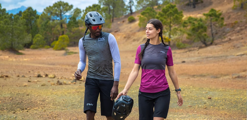 Two cyclists walking in a scenic outdoor setting, wearing cycling gear and carrying helmets.