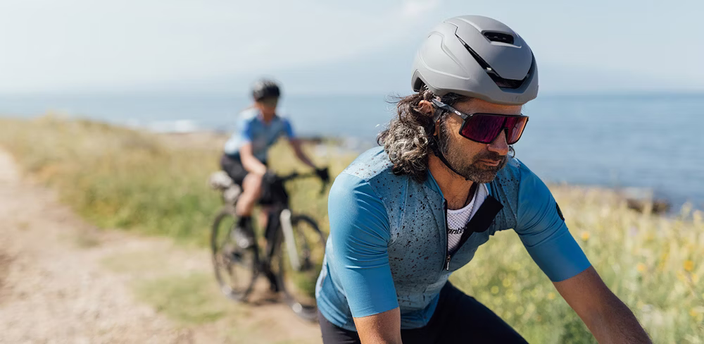 Two cyclists riding on a coastal path, with one in focus wearing a grey helmet and blue cycling jersey.