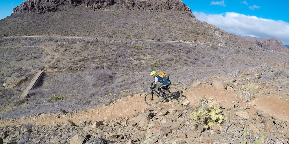 Mountain biker wearing a green helmet and backpack, riding on a rocky desert trail with steep hills and a winding road in the background under a clear blue sky.