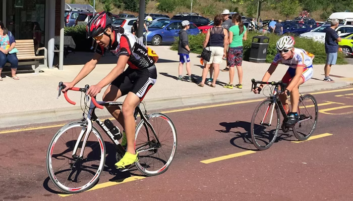 Two cyclists in a road race, with the lead rider wearing a red helmet, black and red jersey, and yellow cycling shoes, followed closely by another cyclist in a white and red jersey. Spectators and parked cars are visible in the background.