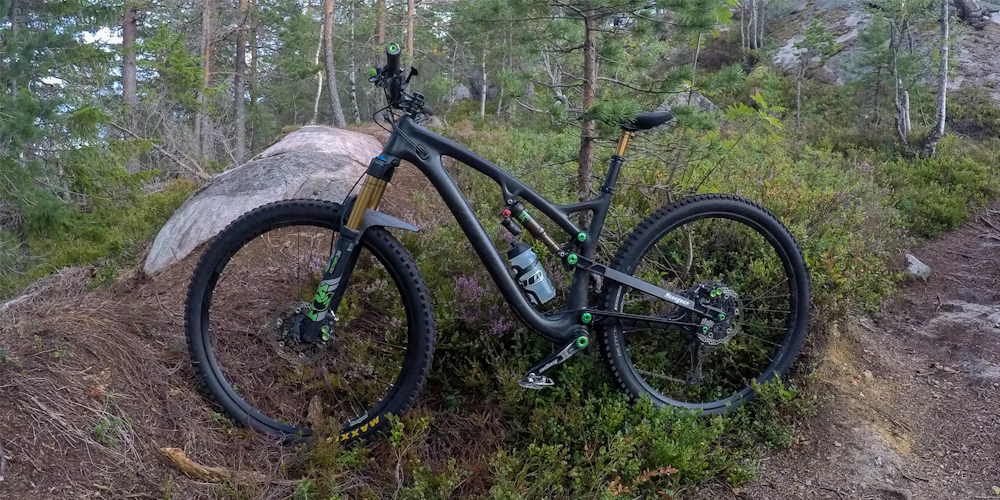 A black mountain bike with green accents resting on a forest trail, surrounded by trees and shrubs.