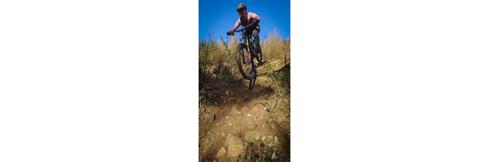 A cyclist performing a downhill jump on a rugged mountain trail, wearing protective gear under a clear blue sky.