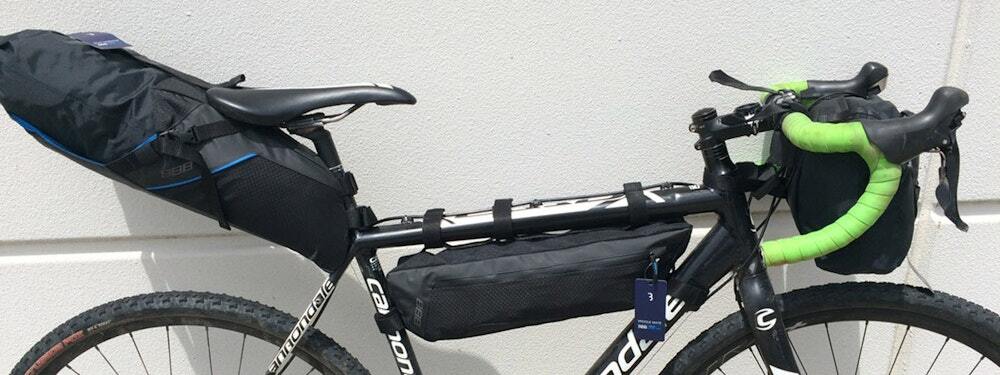 Fully equipped black road bike featuring a seat post bag, frame bag, and handlebar bag for optimal storage, highlighted against a white wall background.