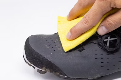 Hand cleaning a black cycling shoe with a yellow sponge, focusing on removing dirt and maintaining the shoe's quality.