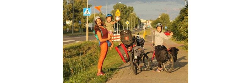 A family on a cycling trip, with a woman holding a child and a man standing beside loaded bicycles on a sunny pathway.