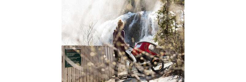 Person standing with a bike trailer in front of a scenic waterfall, surrounded by trees.