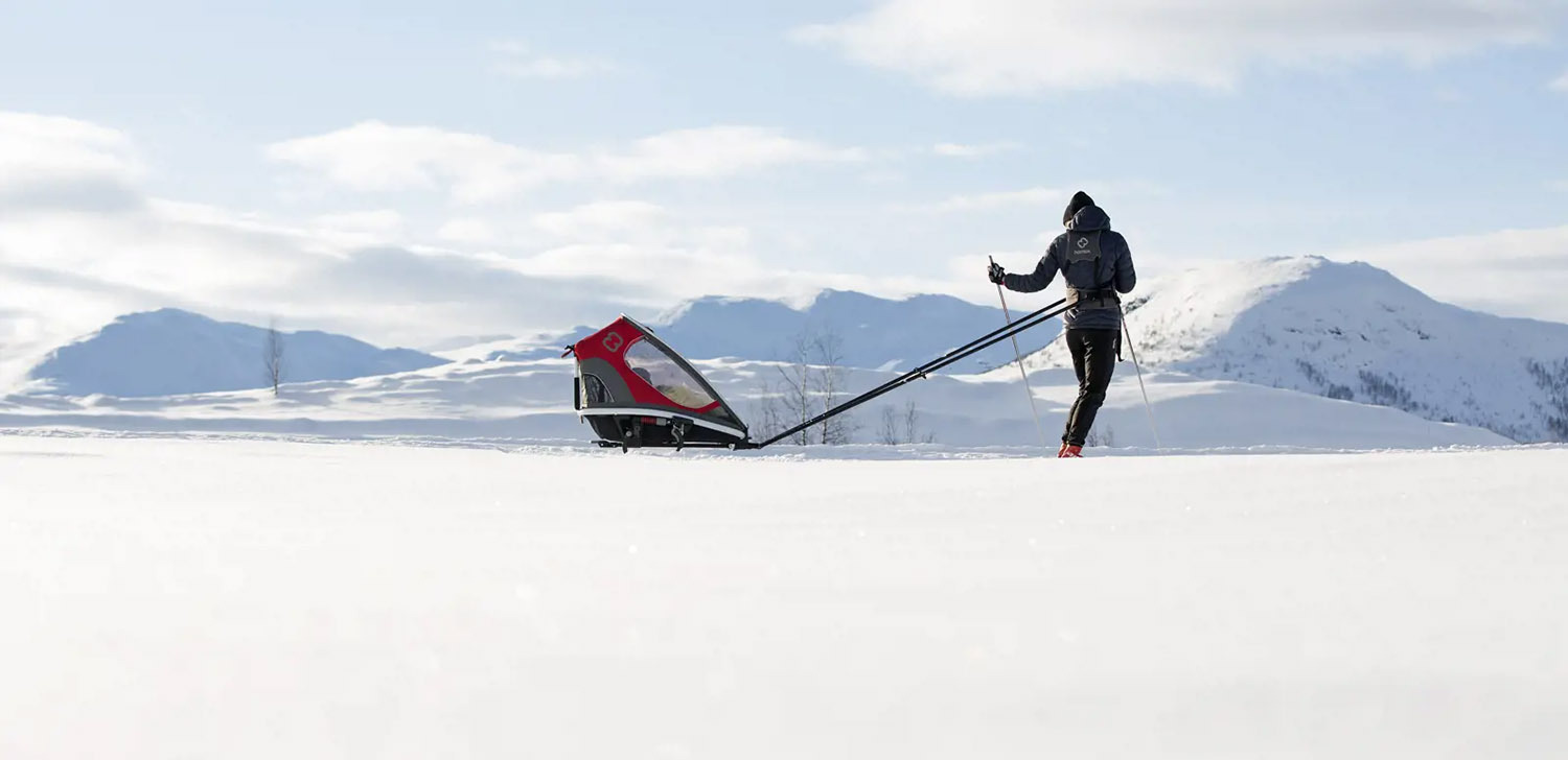 Hamax Outback 2-seat trailer with ski attachment on a snowy winter trail with two passengers