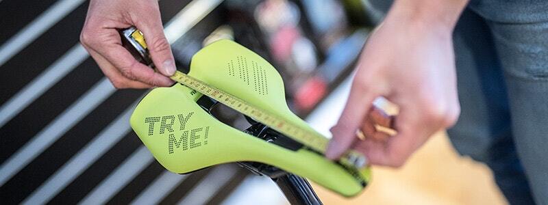 Measuring the width of a bike saddle with 'Try Me!' text printed on it, using a tape measure.