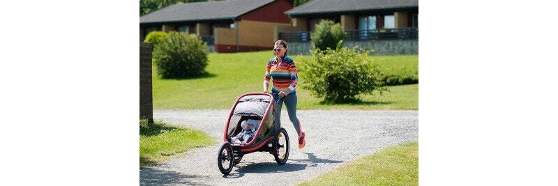 Woman jogging on a path while pushing a child in a three-wheeled stroller, with houses and greenery in the background.