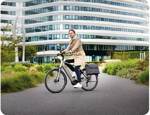 Man in a beige trench coat riding an e-bike on a path surrounded by greenery, with a modern office building in the background.