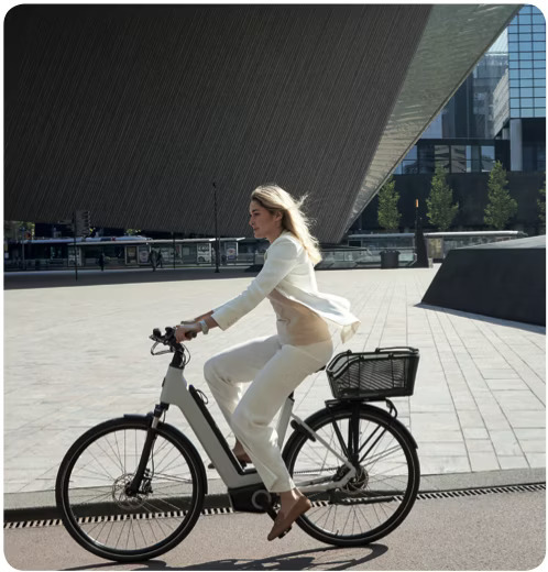 Woman in white outfit riding an e-bike with a front basket in an urban environment with modern architecture in the background.