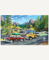  Holiday Traffic wooden jigsaw puzzle shows a line of vintage cars waiting bumper to bumper in traffic down a swerving one-lane road. Pine trees follow along the edge of the mountains in the background. 