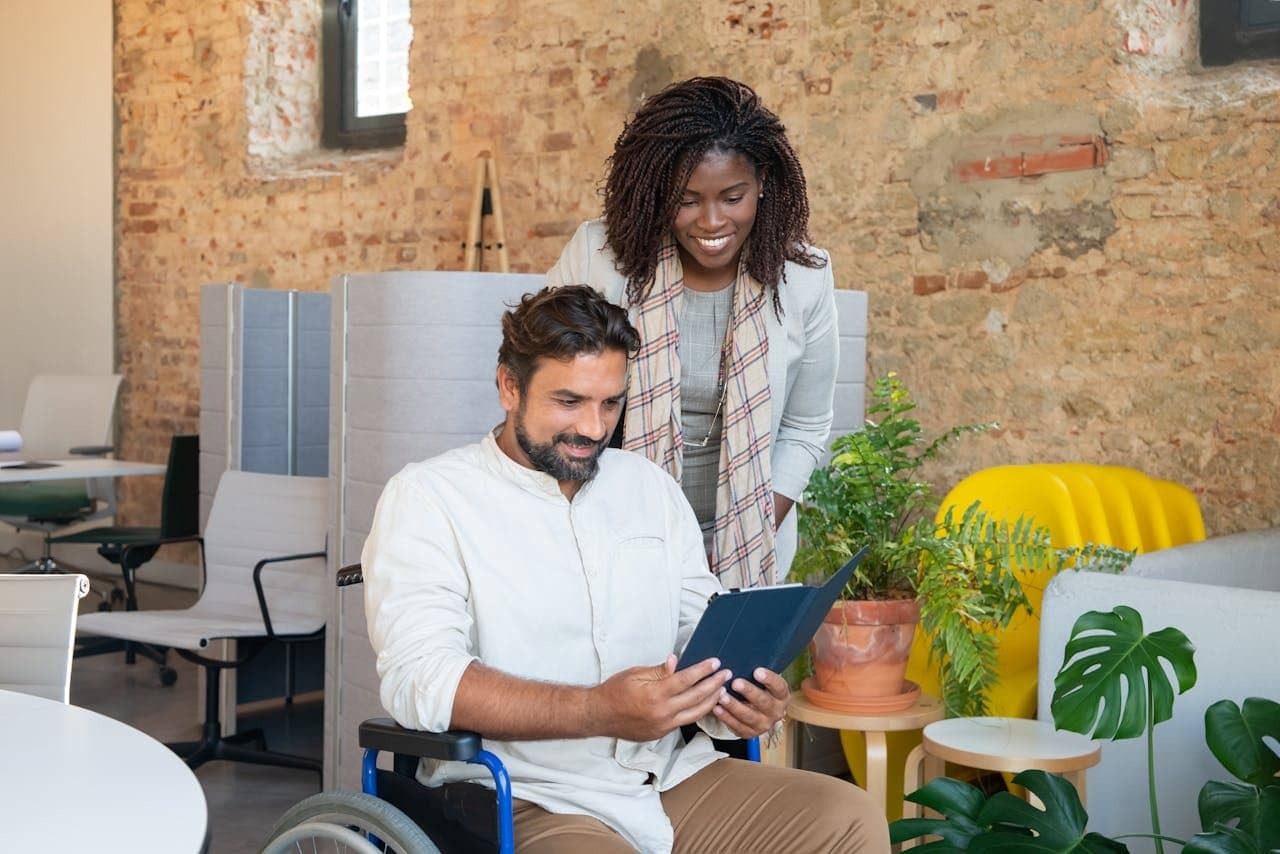 a woman standing beside a man sitting on wheelchair checking a tablet