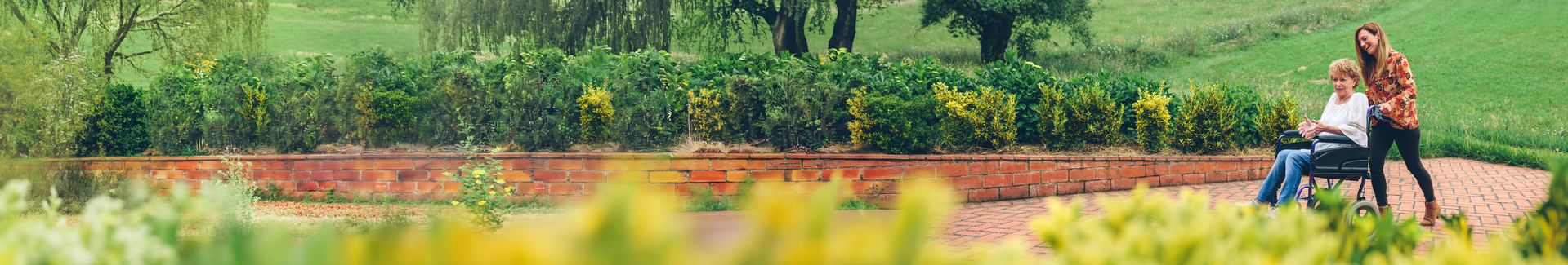 Smiling woman pushing an older woman in a wheelchair along a brick garden path