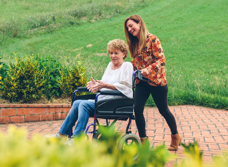 Smiling woman pushing an older woman in a wheelchair along a brick garden path