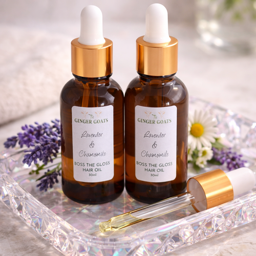 Two amber glass dropper bottles of Lavender and Chamomile Hair Oil from The Ginger Goats, displayed on a crystal-style tray with a pipette, lavender sprigs and chamomile flowers in a soft, calming setting.