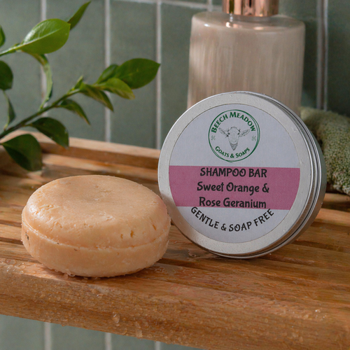 Round Sweet Orange and Rose Geranium shampoo bar and matching tin from Beech Meadow Goats and Soaps displayed on a wooden shower shelf with soft green tiles in the background, showing a natural handmade hair care product in a calm bathroom setting.