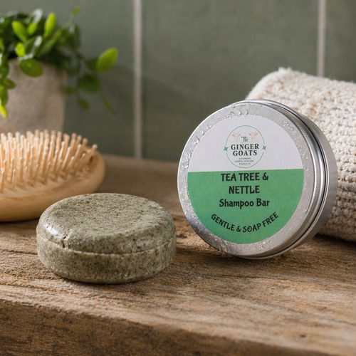 Tea Tree and Nettle shampoo bar beside its metal tin packaging on a rustic wooden surface in a softly lit bathroom setting, with a wooden hairbrush and folded towel in the background.