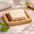 Rectangular beech wooden soap dish with slatted drainage design holding a handmade soap bar on a marble bathroom surface.